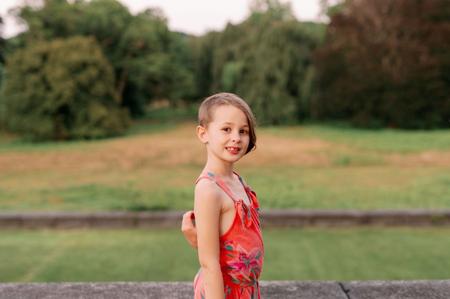 Child in red dress poses outdoors on grassy hillside during summer photoshoot.