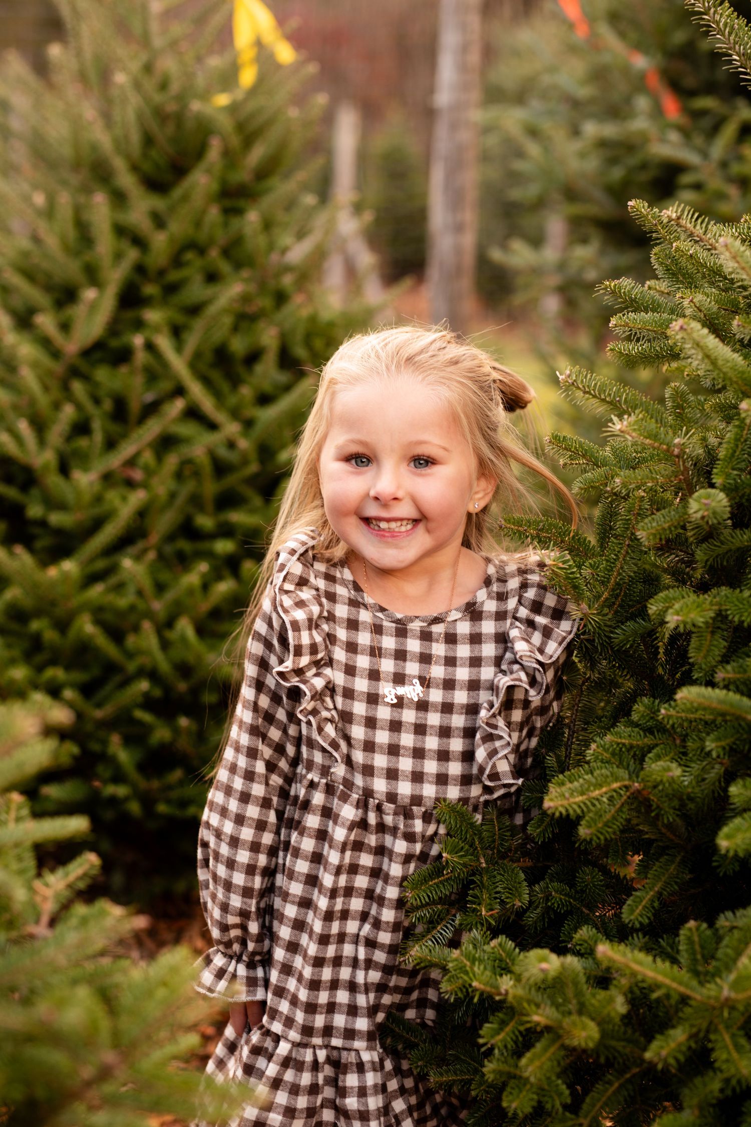 A child in a checkered dress smiles joyfully while standing among evergreen Christmas trees at a tree farm.