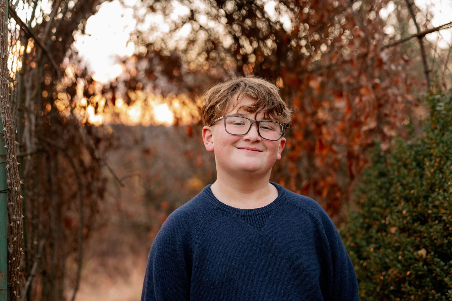 A person in a navy sweater and glasses standing in front of autumn foliage at sunset.