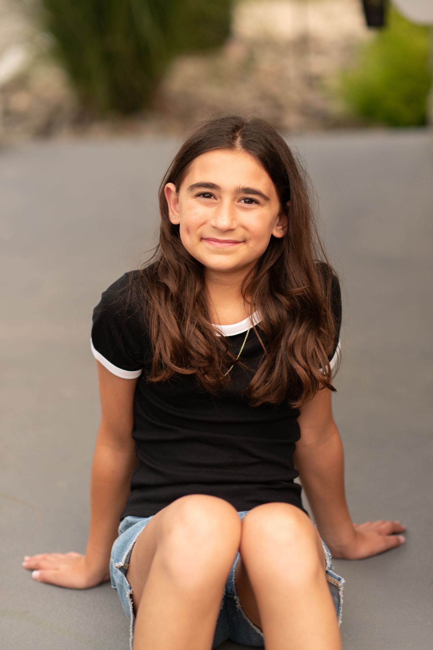 Young person in a black t-shirt and denim shorts sitting casually outdoors on a sunny day.