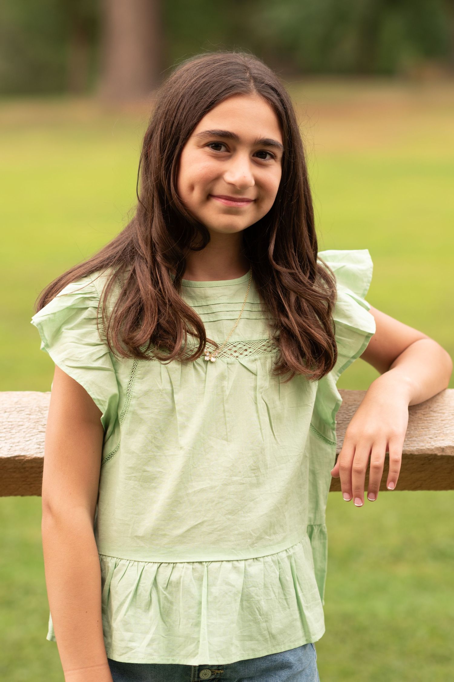A person in a light green ruffled top leans on a wooden railing in an outdoor setting with grass in the background.