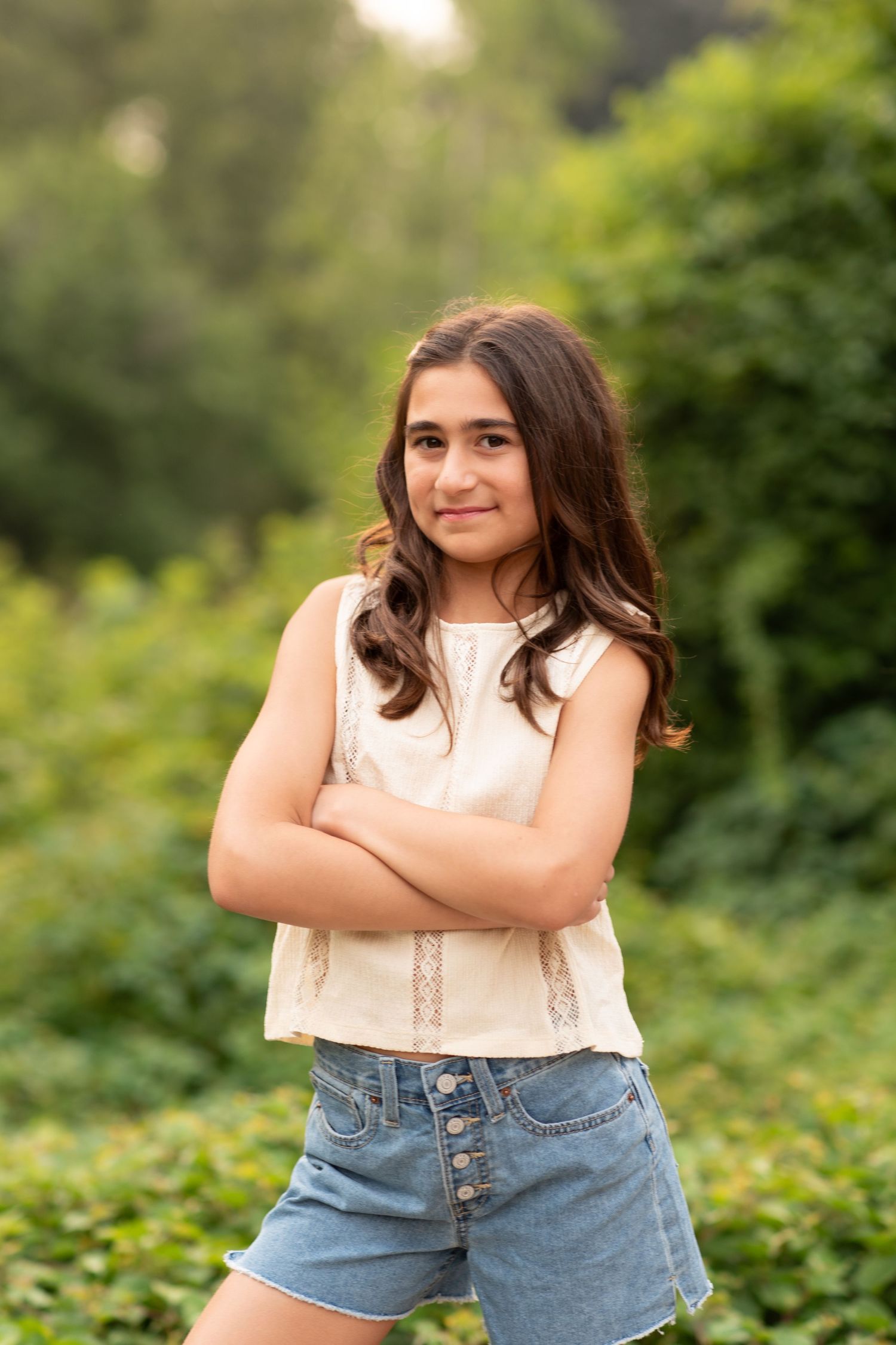 A person poses outdoors in a white sleeveless top and denim shorts against a green natural background.