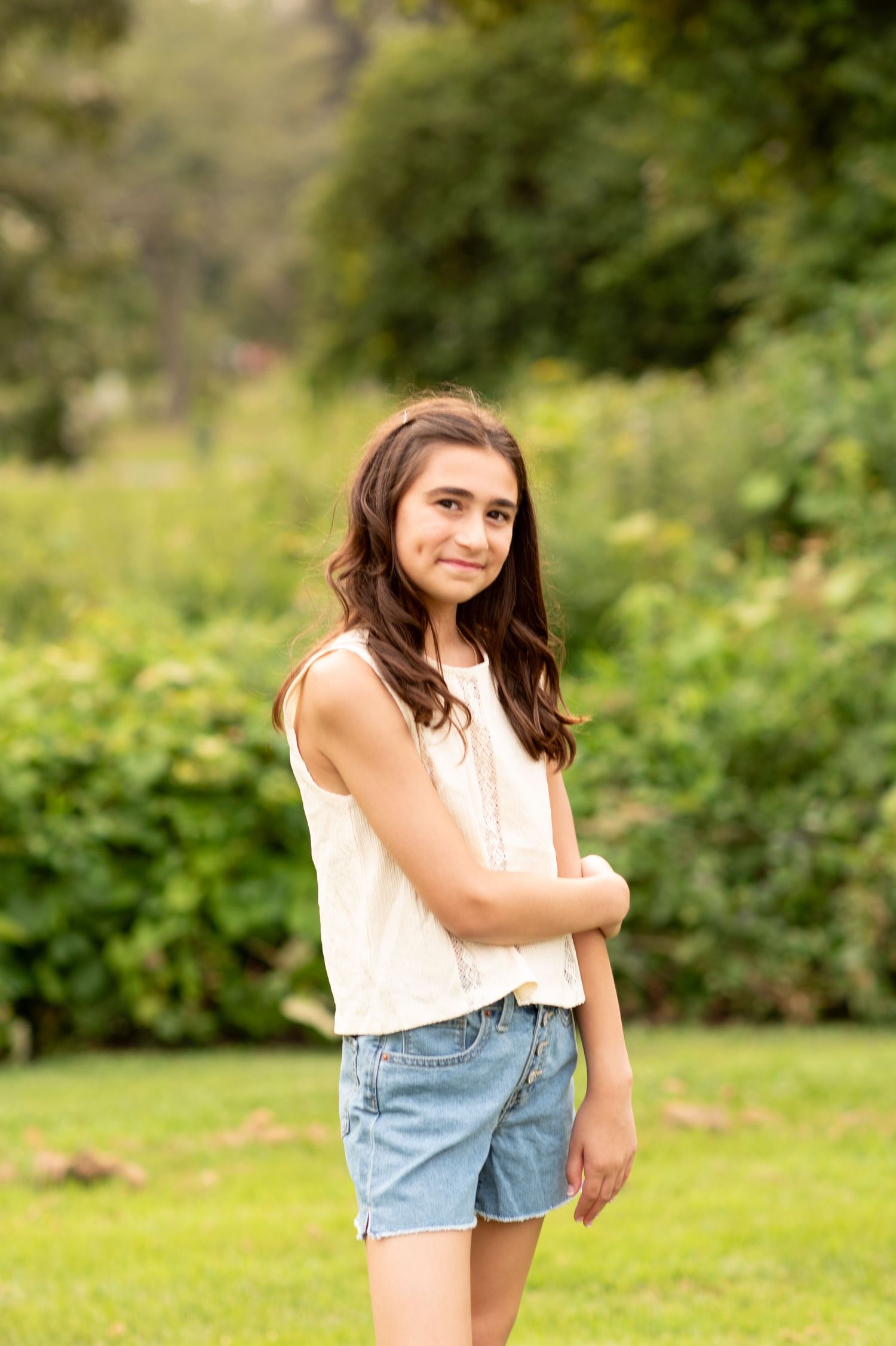 Person wearing a white top and denim shorts poses casually in a lush green outdoor setting on a sunny day.