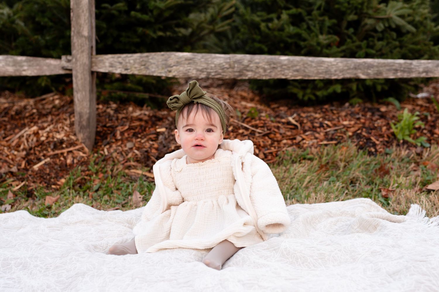 Small child wearing a cream-colored dress sits on a white blanket against a rustic wooden fence and mulch background.