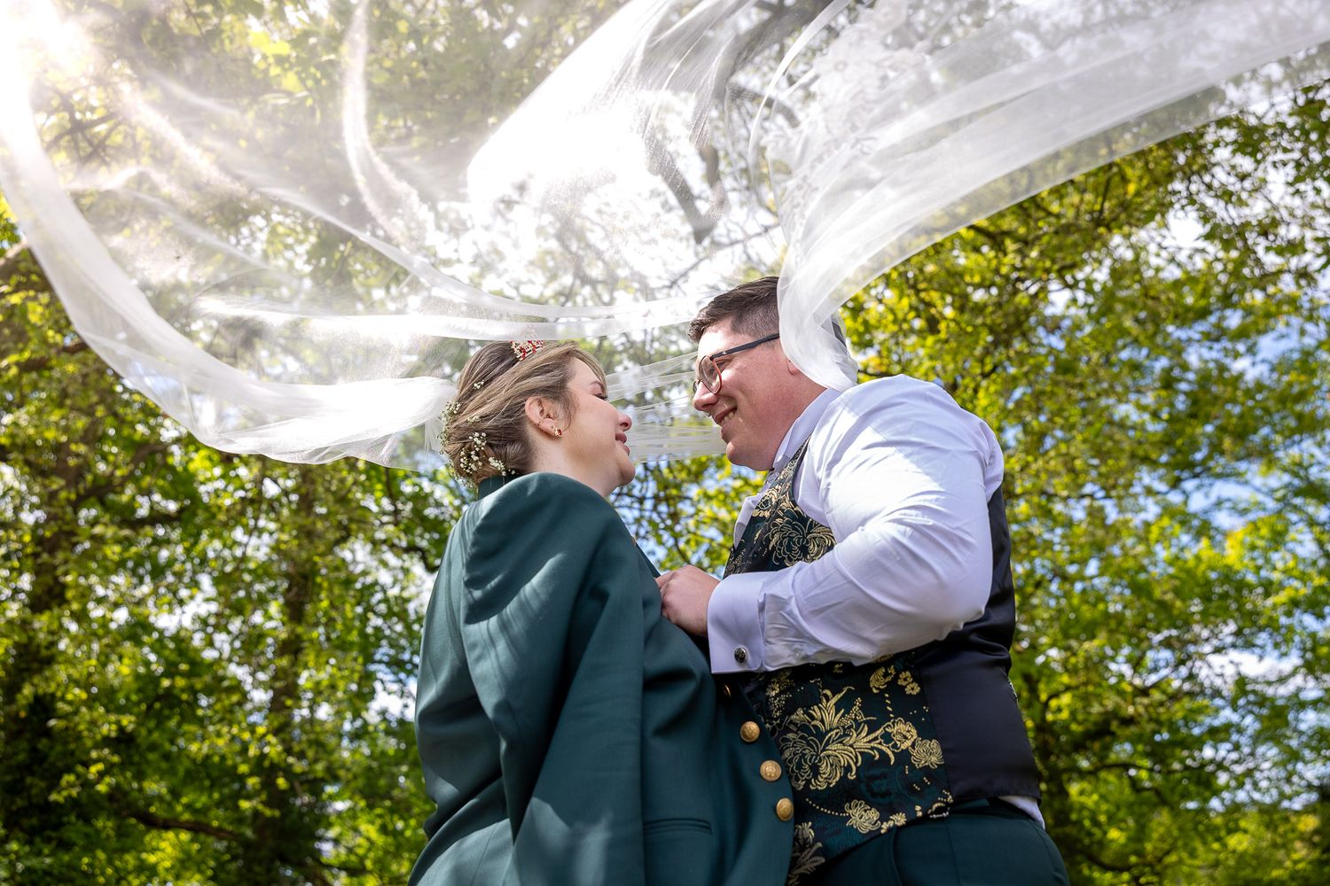 Wedding veil flowing dramatically in the breeze against a backdrop of green trees.