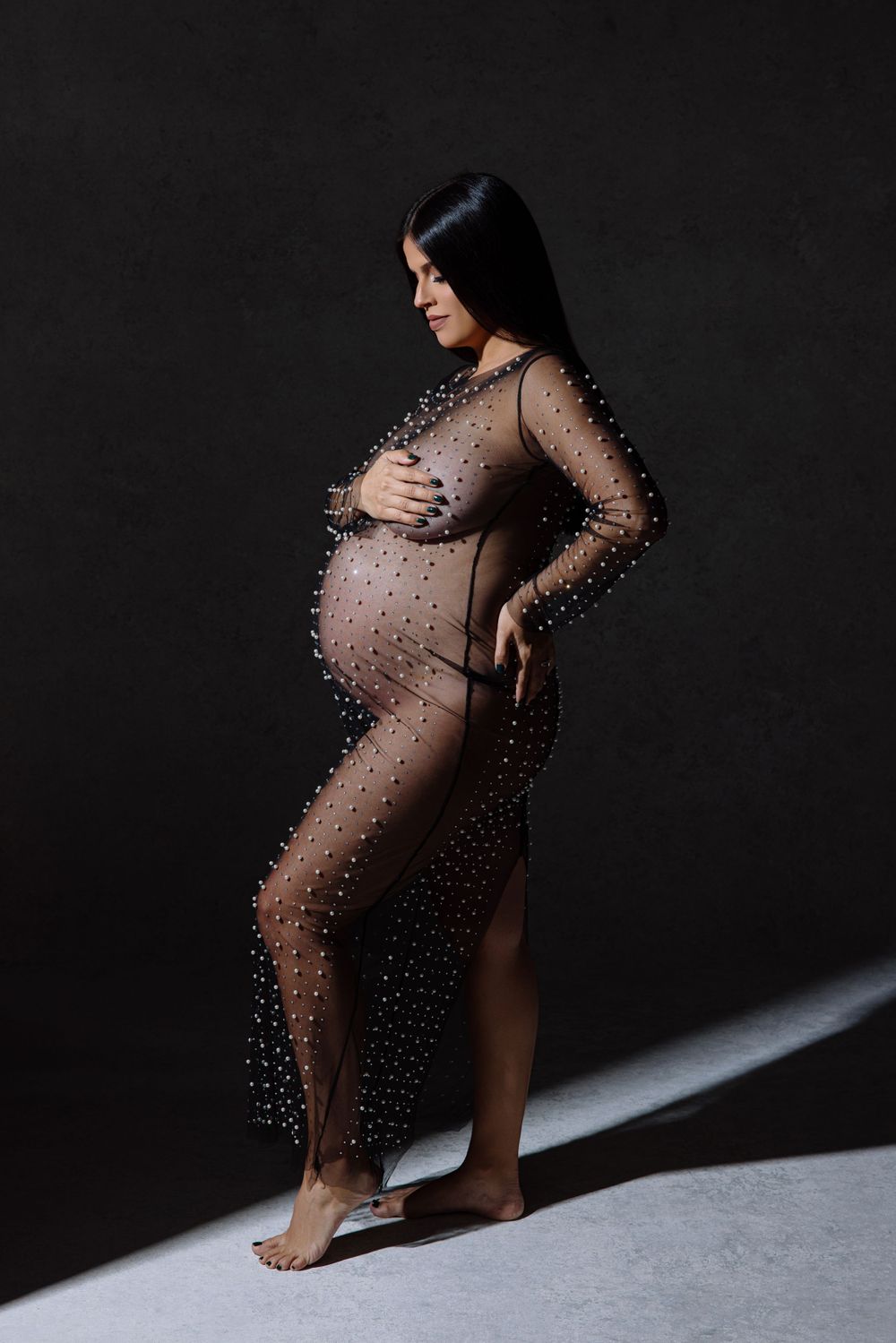 An artistic maternity photo of a silhouette in a sheer beaded dress against a dark background with dramatic lighting.