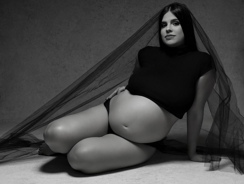 Black and white artistic maternity photo showing a pregnant figure in dark top reclining with flowing veil.
