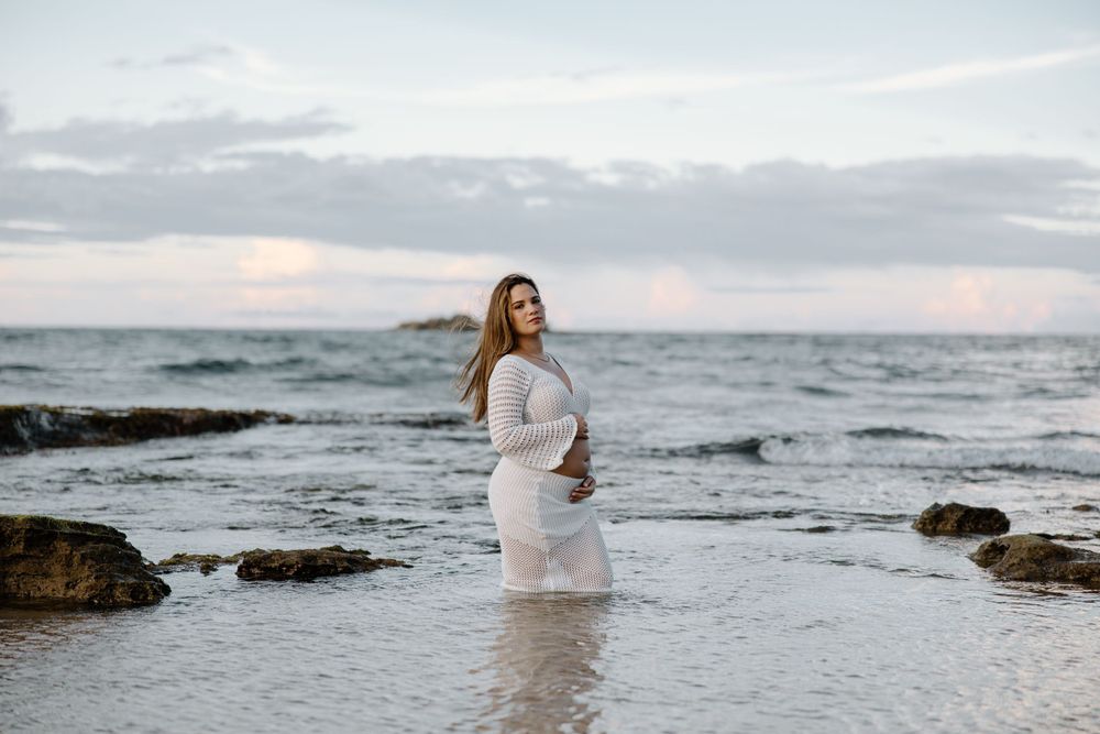 A person in white dress standing in shallow ocean water during sunset.