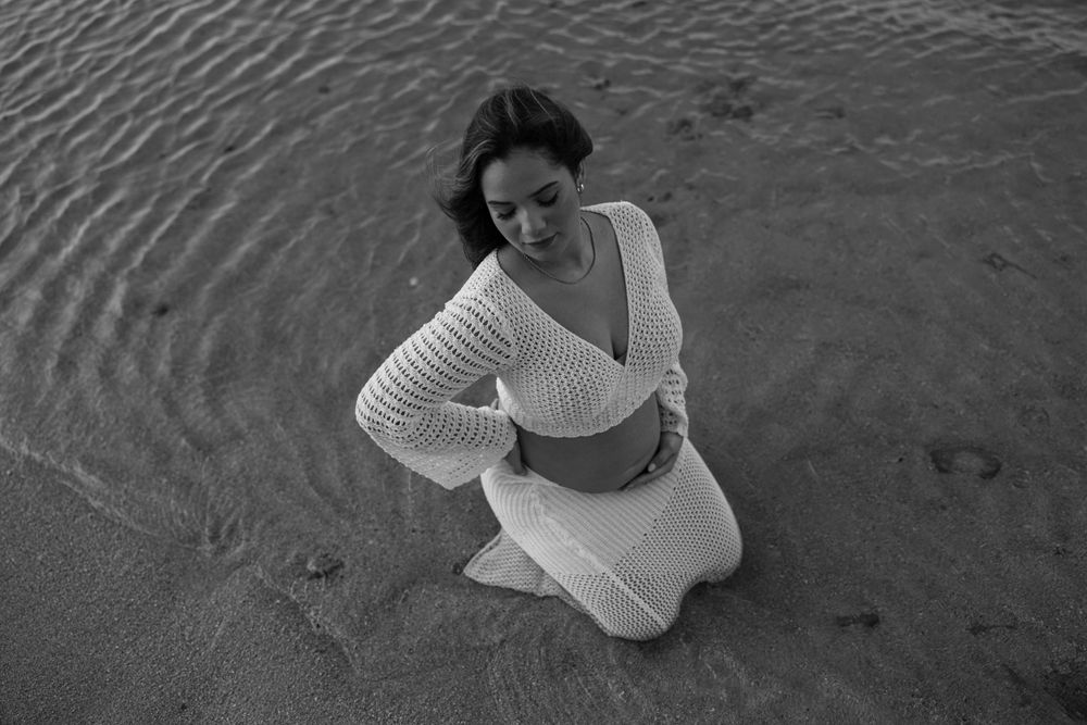 A person in white clothing kneeling on wet sand at the beach.