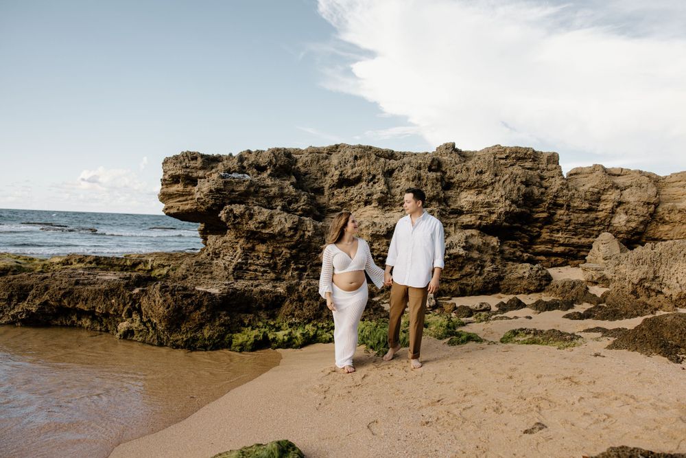 A couple in white outfits standing on a beach near rocky cliffs during golden hour.