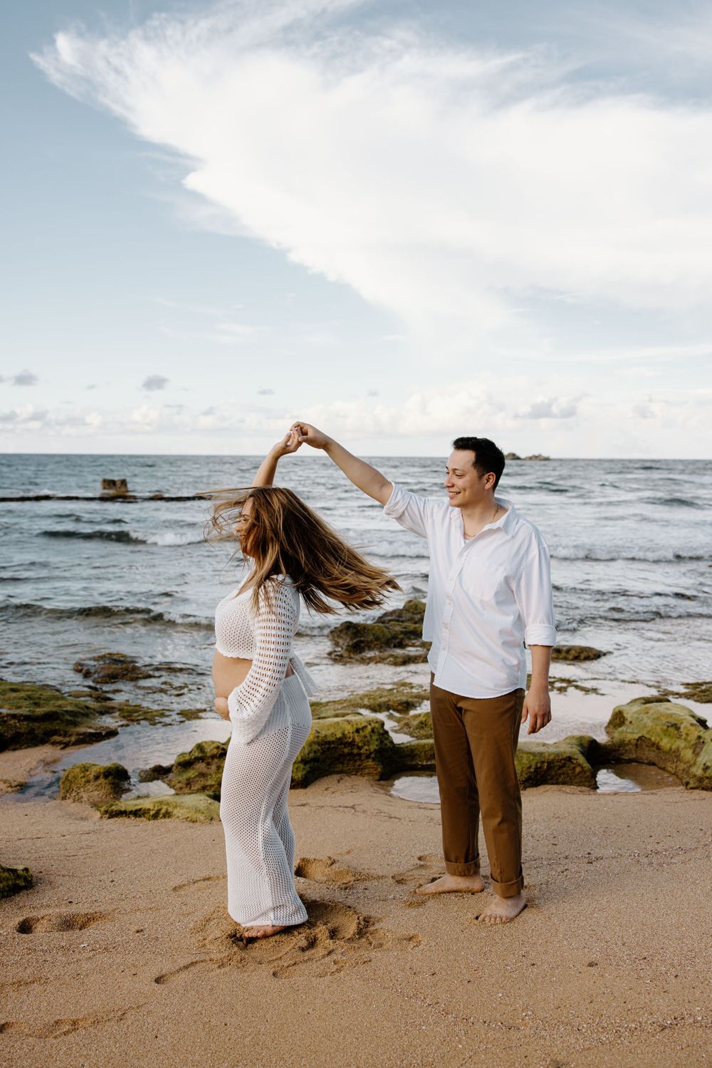 A couple dancing together on a scenic beach with waves and rocks in the background.