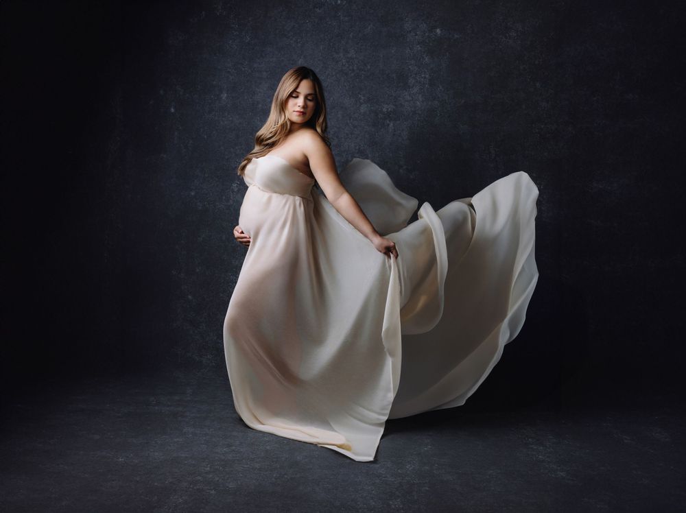 Artistic maternity photo with flowing white dress against dark background.