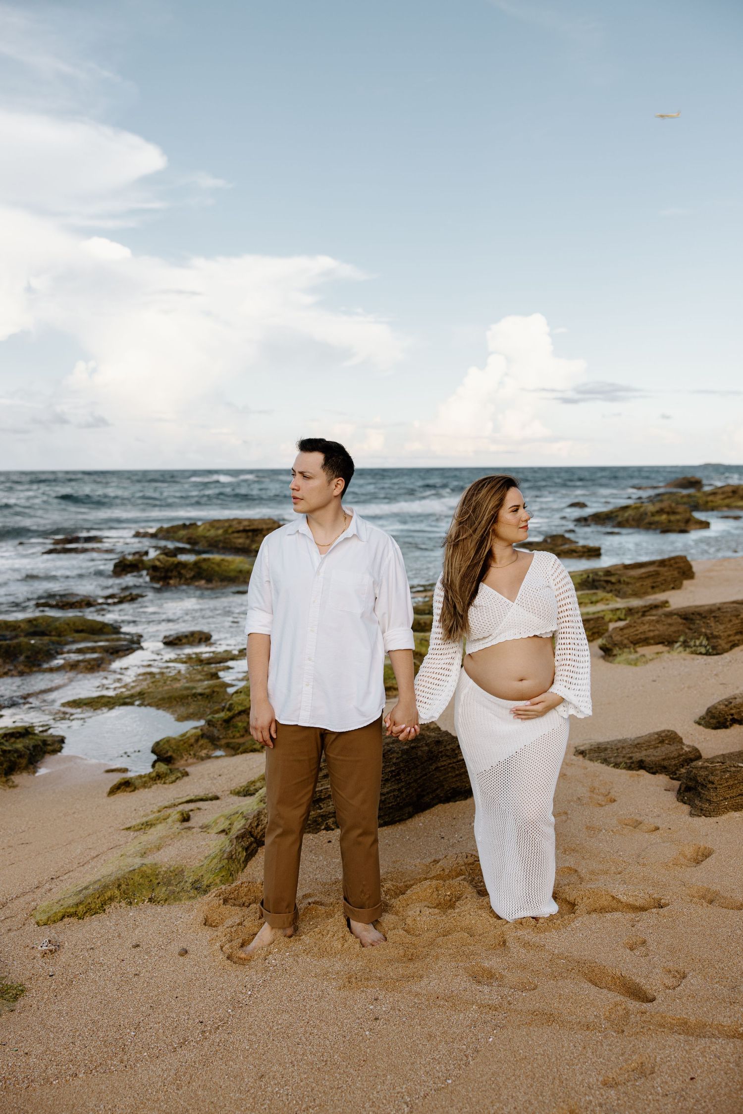 Expecting couple holding hands on beach with ocean and cloudy sky backdrop.
