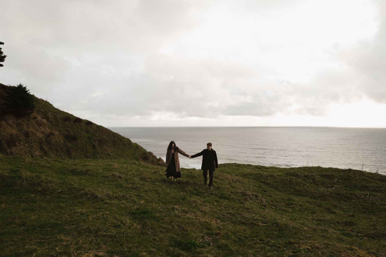 Two figures hold hands while walking along a scenic coastal cliff at dusk.