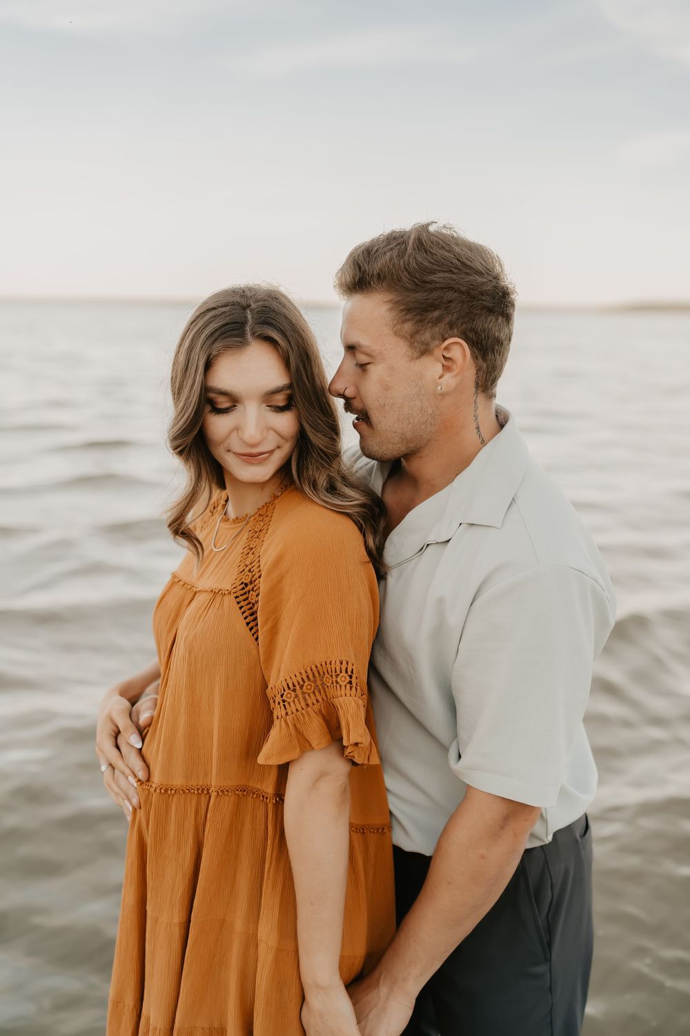 Romantic couple embraces on beach during sunset while wearing orange dress and grey shirt.