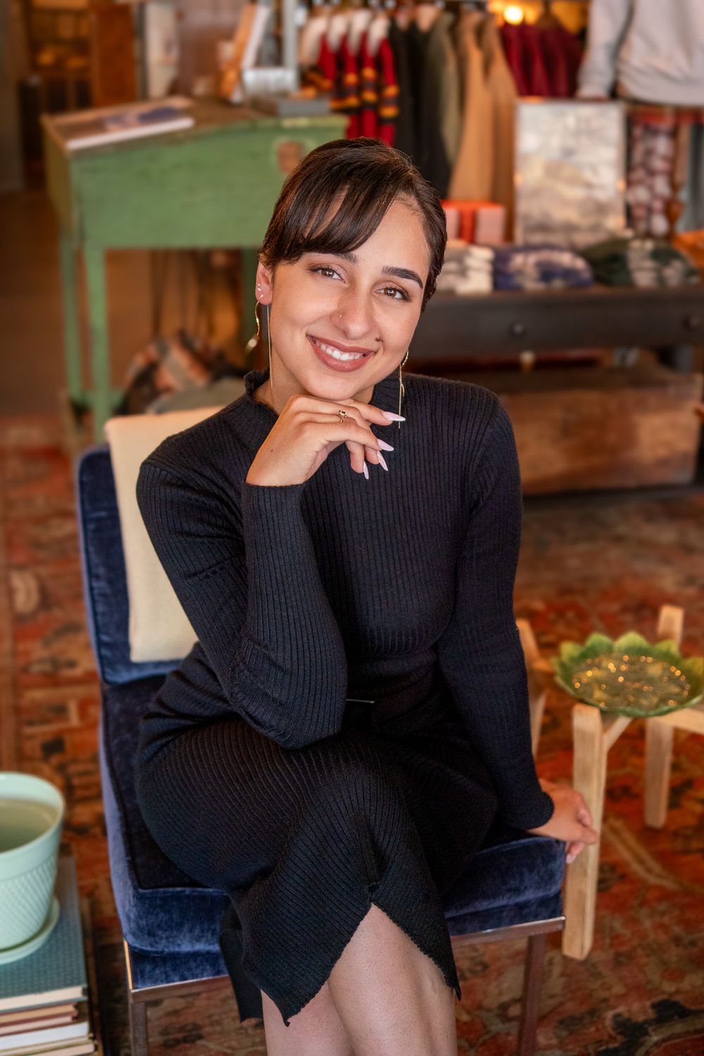 A person in a black ribbed sweater dress sits in a blue velvet chair in a cozy retail setting.