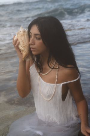A dreamy beach portrait featuring someone in white holding a seashell while wearing pearl necklaces in the ocean.