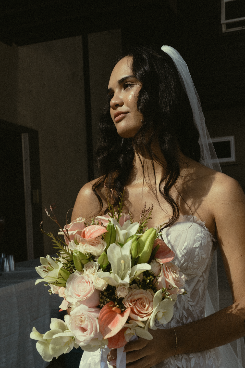 A bride in a white strapless gown holds a beautiful bouquet of pink roses and white lilies in natural lighting.