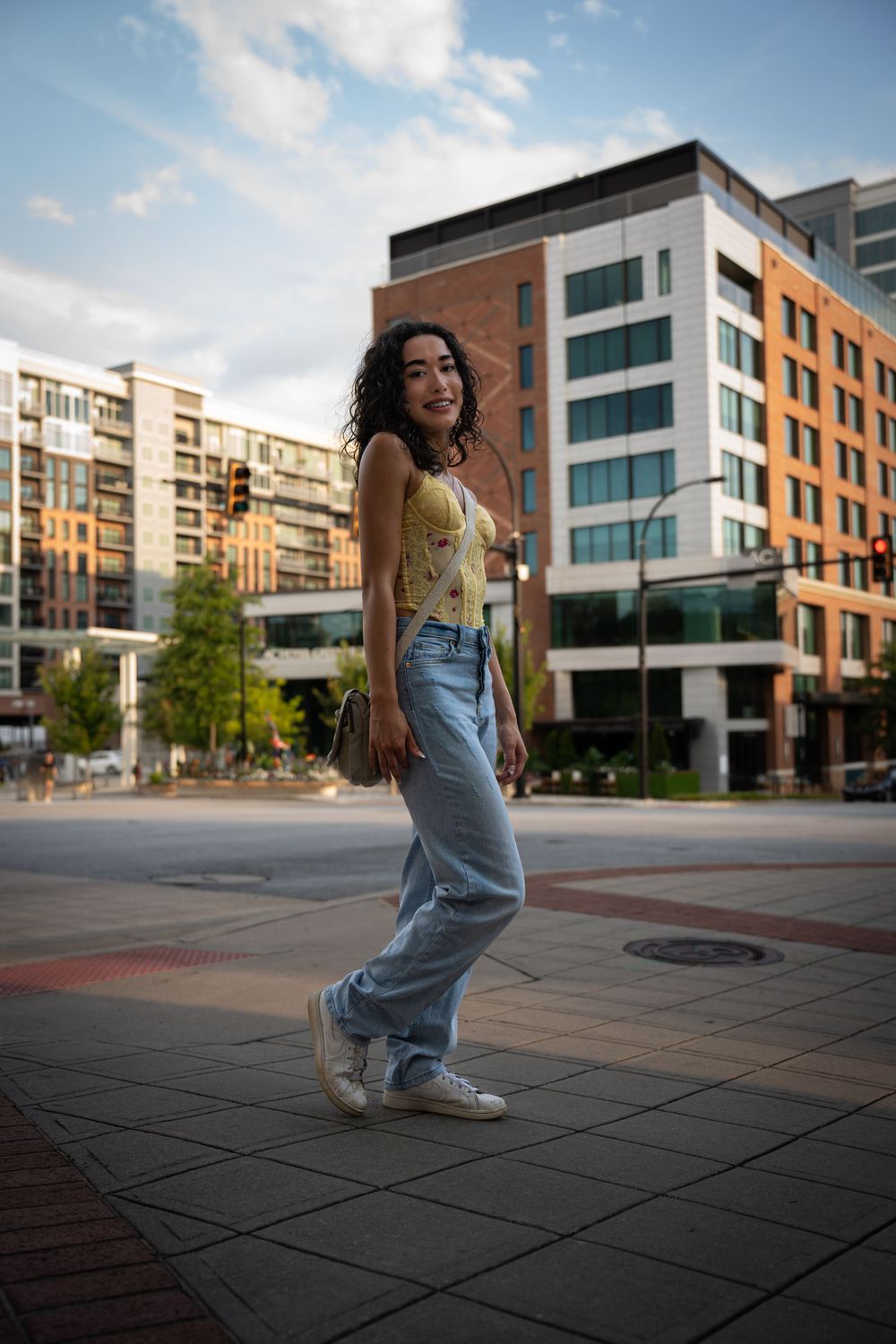 Stylish woman in yellow top and baggy jeans poses on urban plaza with modern apartment buildings in background.