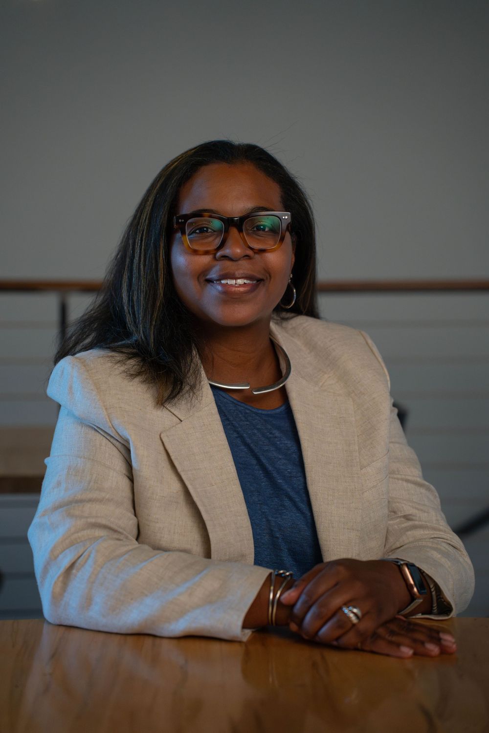 Professional wearing a beige blazer and navy top smiles warmly while seated at a desk in an office setting.