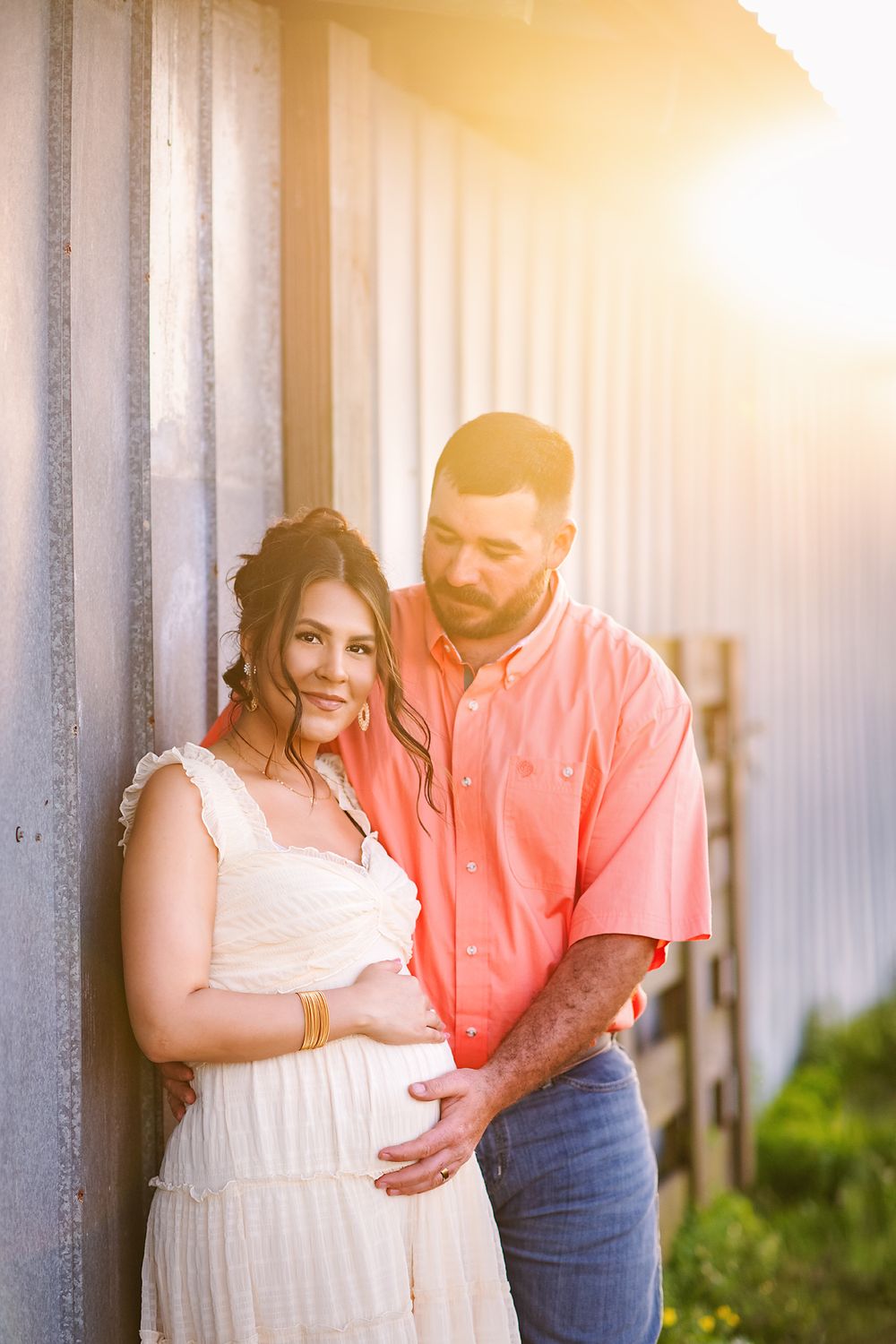 Expecting couple poses against metal wall in golden sunset light, man in coral shirt embracing pregnant woman.