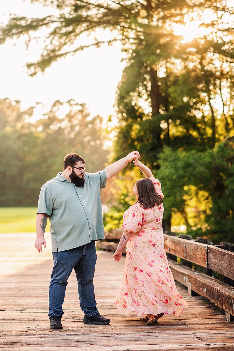 Couple dancing on a wooden bridge at golden hour, man in green shirt twirling woman in floral dress outdoors.