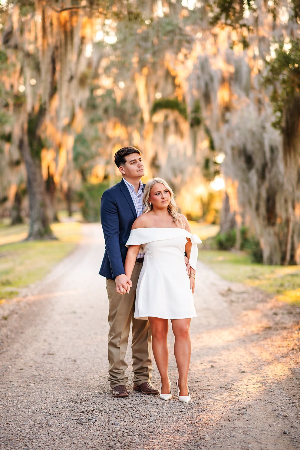 Engaged couple holding hands on a tree-lined gravel path at golden hour sunset.