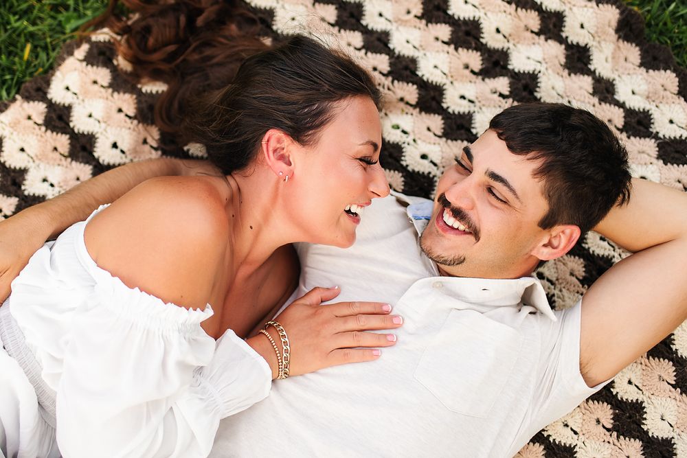 Couple laughing and lying on a checkered blanket outdoors, woman in white dress, man in white shirt.