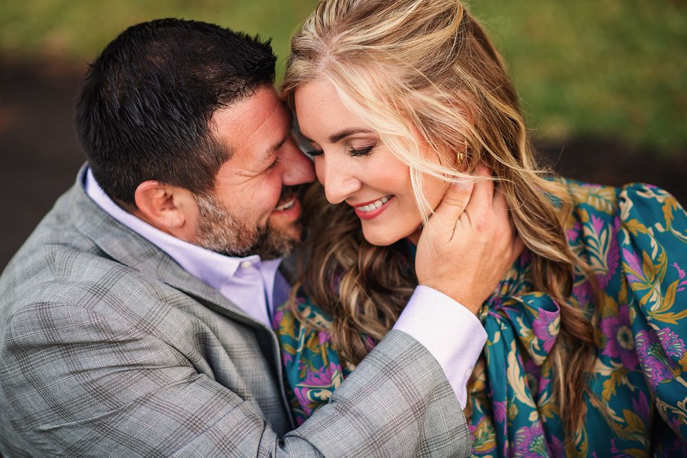 A couple shares a tender romantic moment outdoors, the man in a gray suit touching the womans face gently.