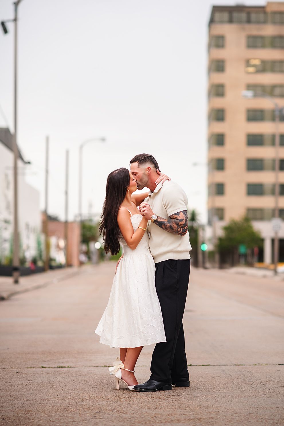 Couple shares romantic kiss while standing in middle of urban street in casual elegant attire.