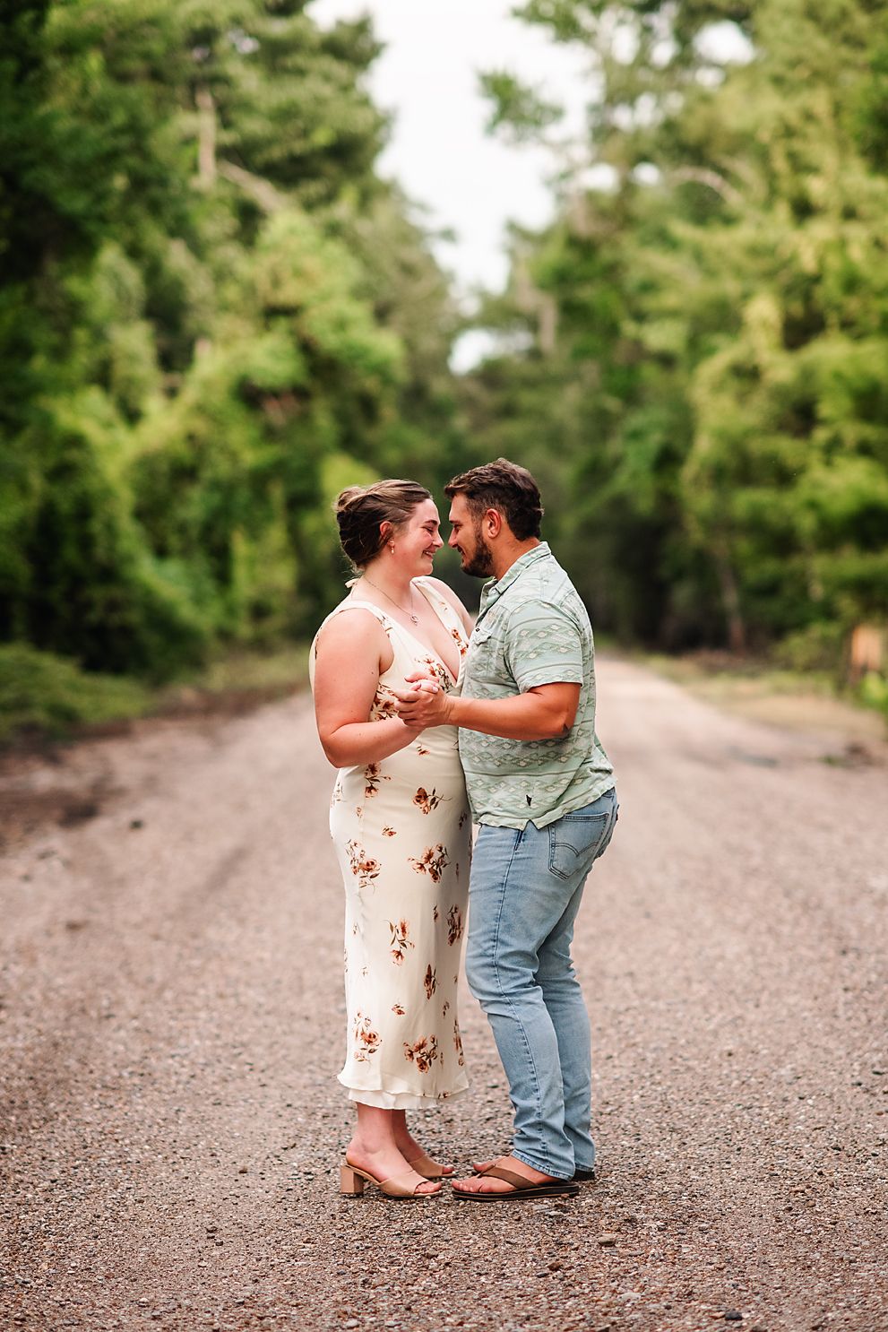 Couple holding hands and smiling at each other on a tree-lined gravel road.