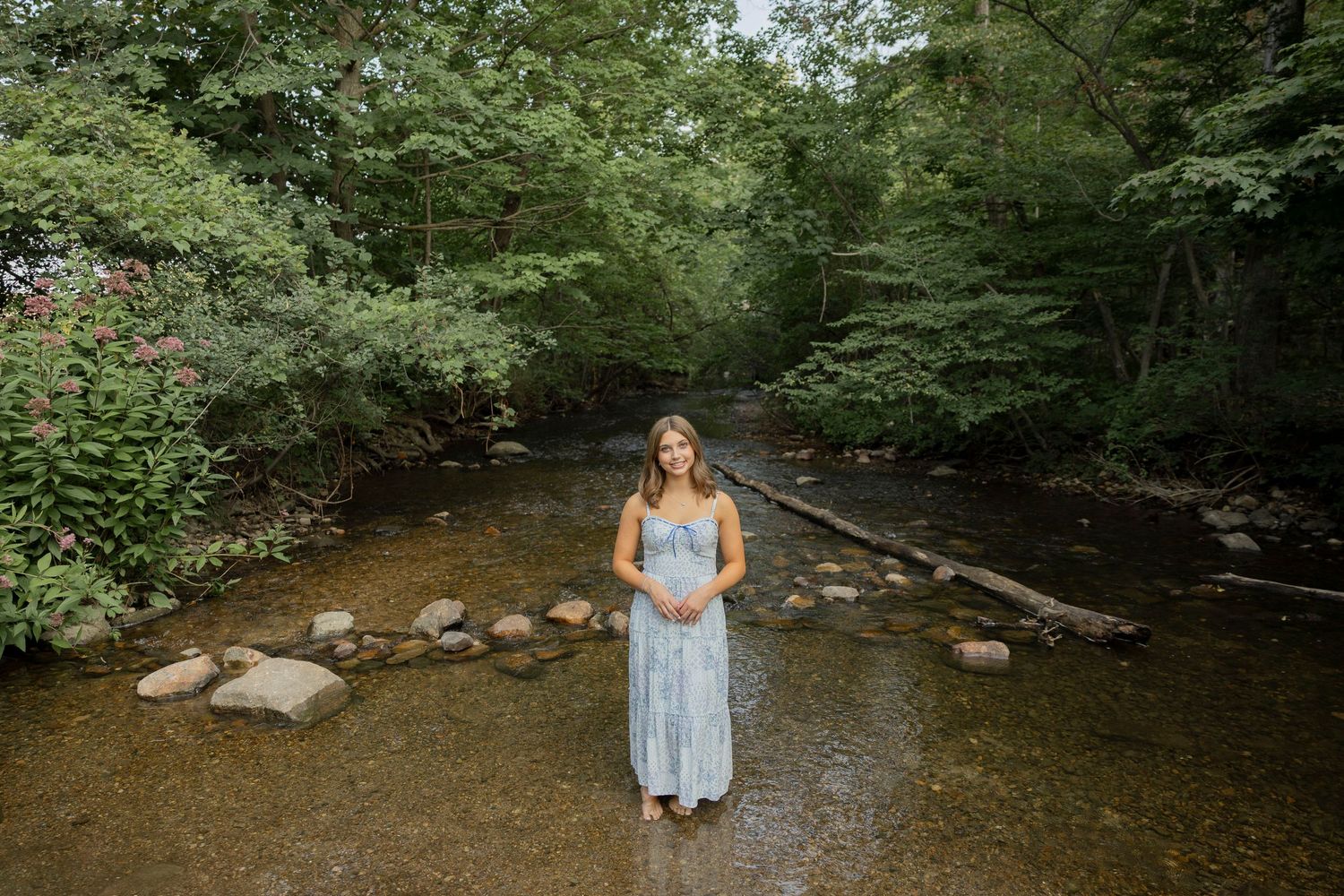 high school senior in blue maxi dress standing in a shallow creek surrounded by lush green forest trees.