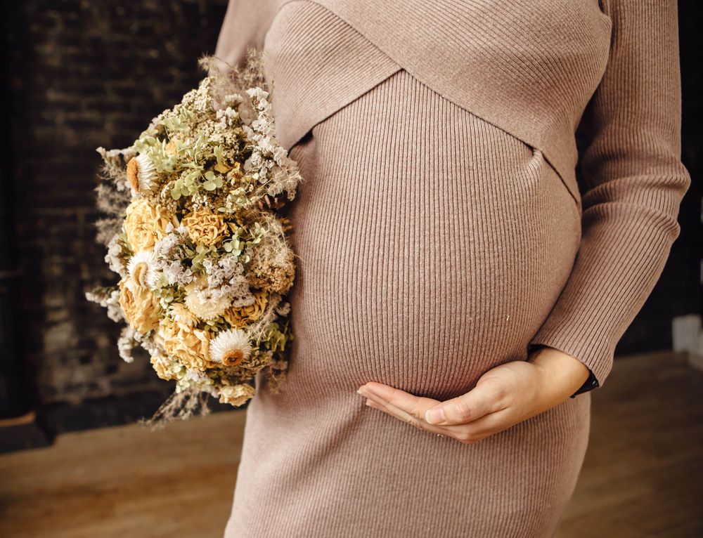 Close up of a pregnant belly in a beige ribbed sweater dress with dried flowers being held to the side.