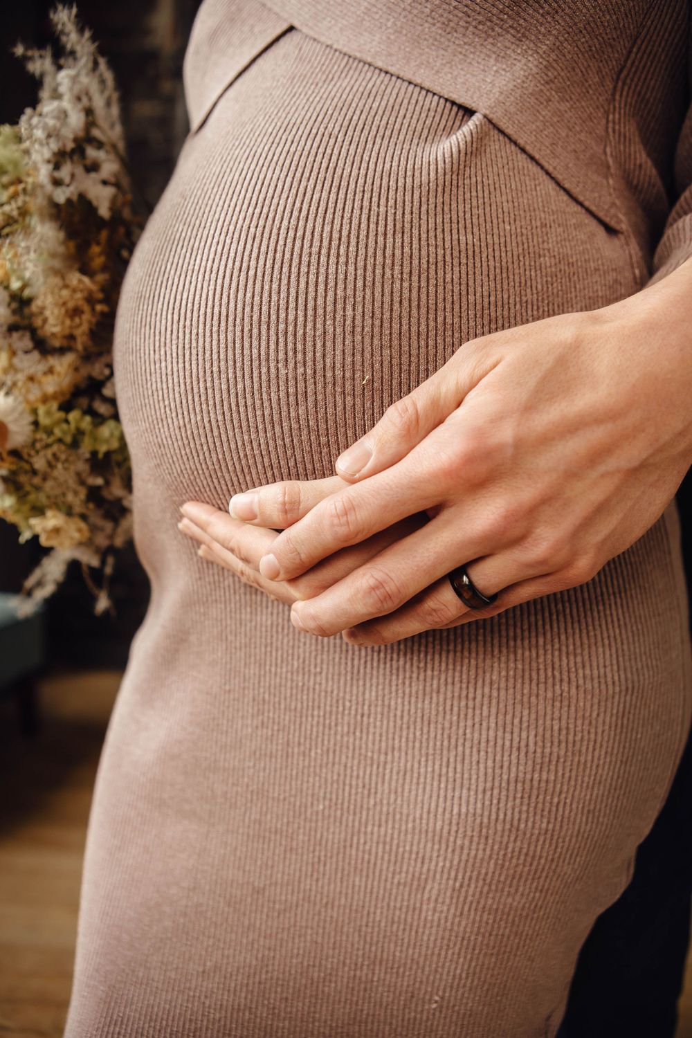Close-up of a pregnant belly in a brown ribbed dress with hands gently resting on the bump.