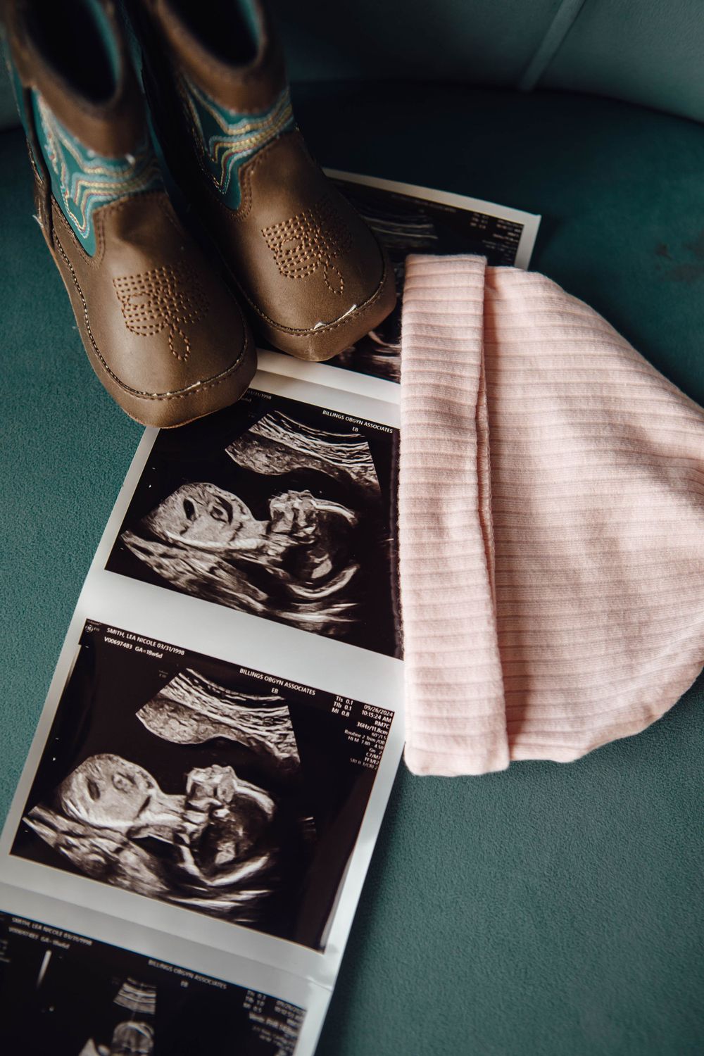 Ultrasound sonogram images displayed next to small brown cowboy boots and a pink baby hat on a dark green surface.