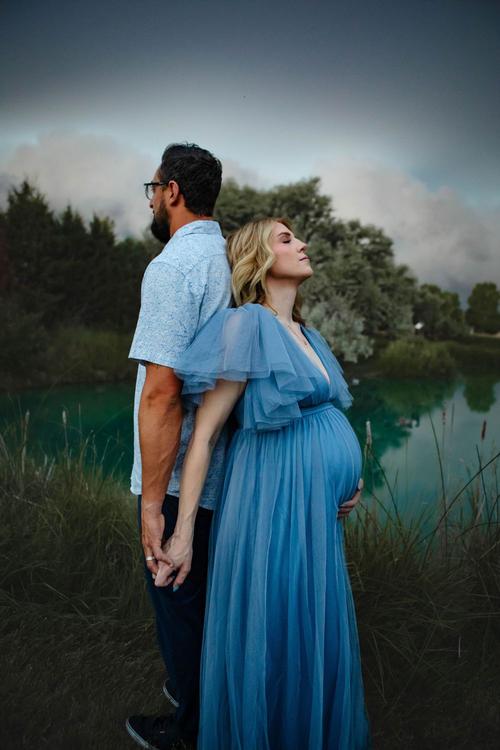 A couple stands back-to-back by a lake at sunset, with the woman wearing a flowing blue maternity gown.