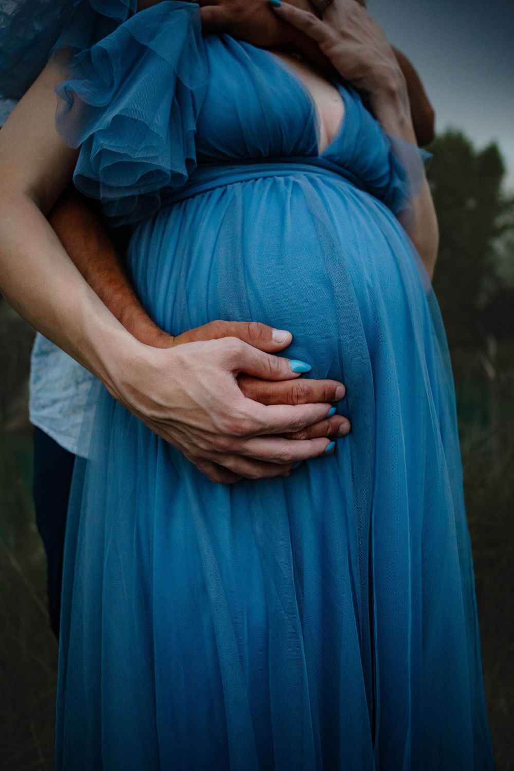 Hands gently embrace a pregnant belly draped in flowing turquoise blue chiffon dress against a moody outdoor backdrop.