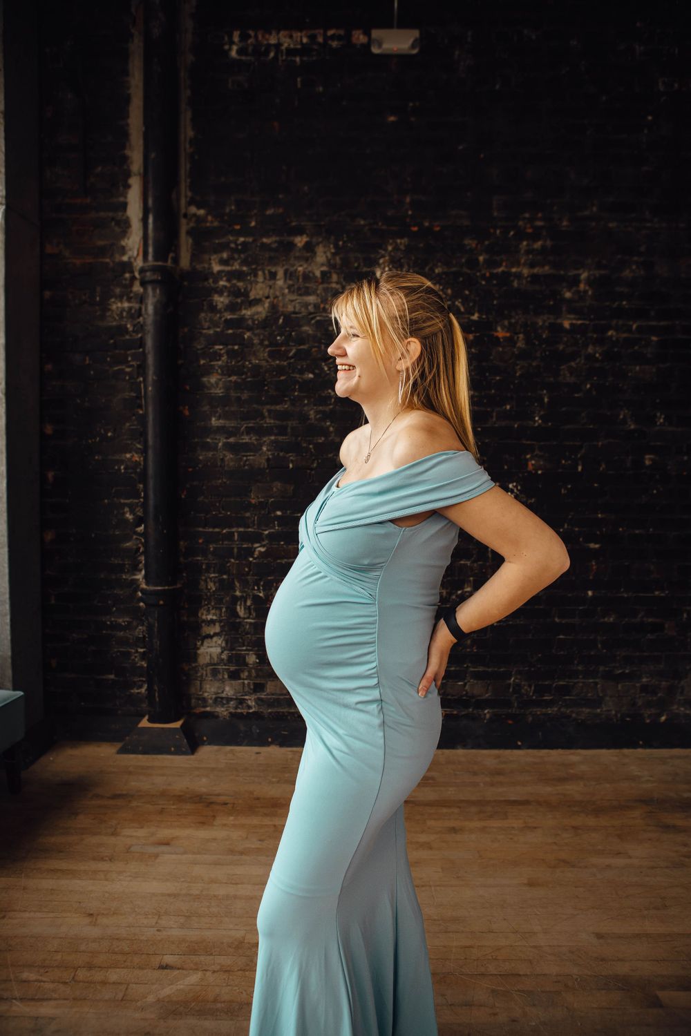 A pregnant woman in a light blue form-fitting dress poses against a dark brick wall in an indoor setting.