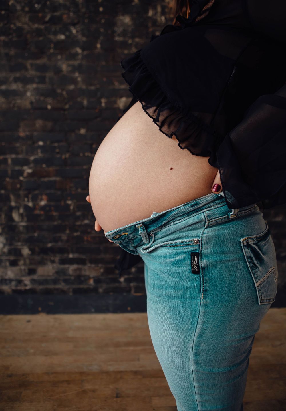 A pregnant woman's bare belly shown against dark jeans and a black top with brick wall backdrop.
