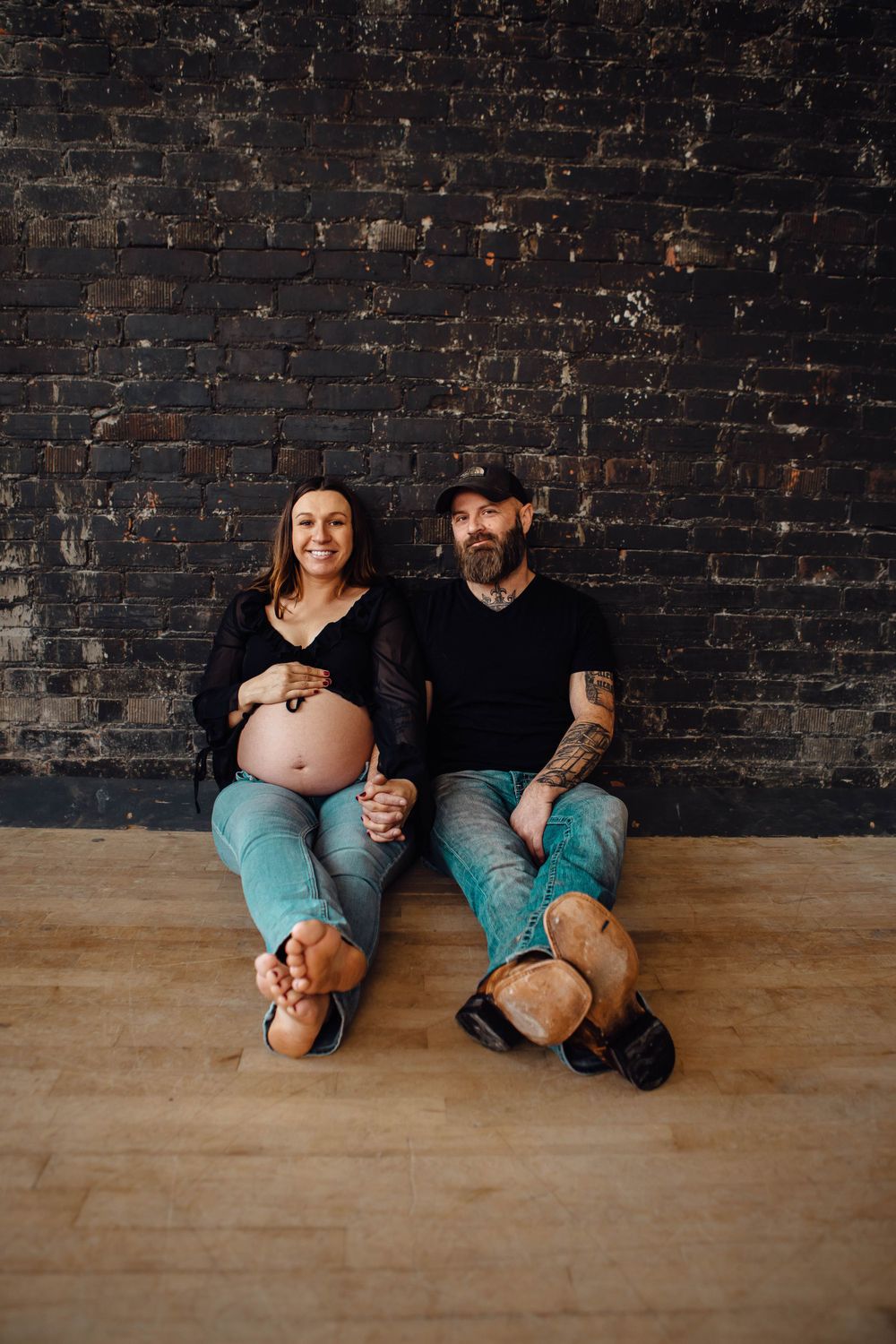 Expectant couple sits together on wooden floor against dark brick wall wearing casual denim and black clothing.