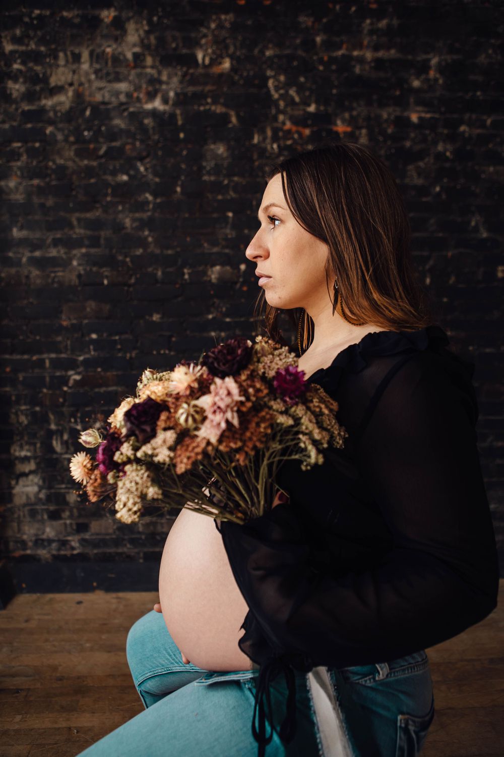 A pregnant woman in a black sweater and jeans holds a vibrant bouquet against a dark brick wall background.
