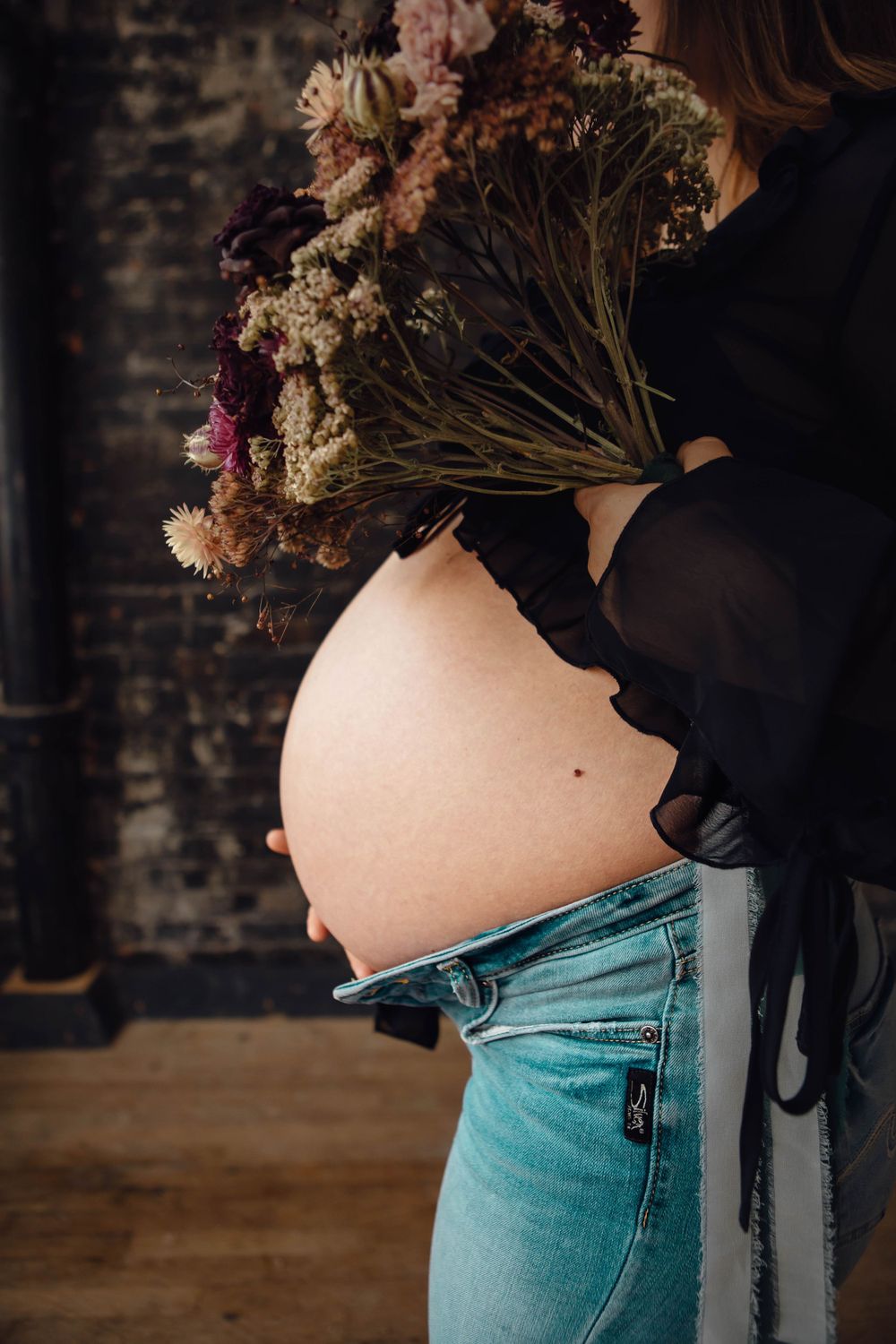 A pregnant woman in blue jeans holds dried flowers above her bare belly in a moody indoor setting.