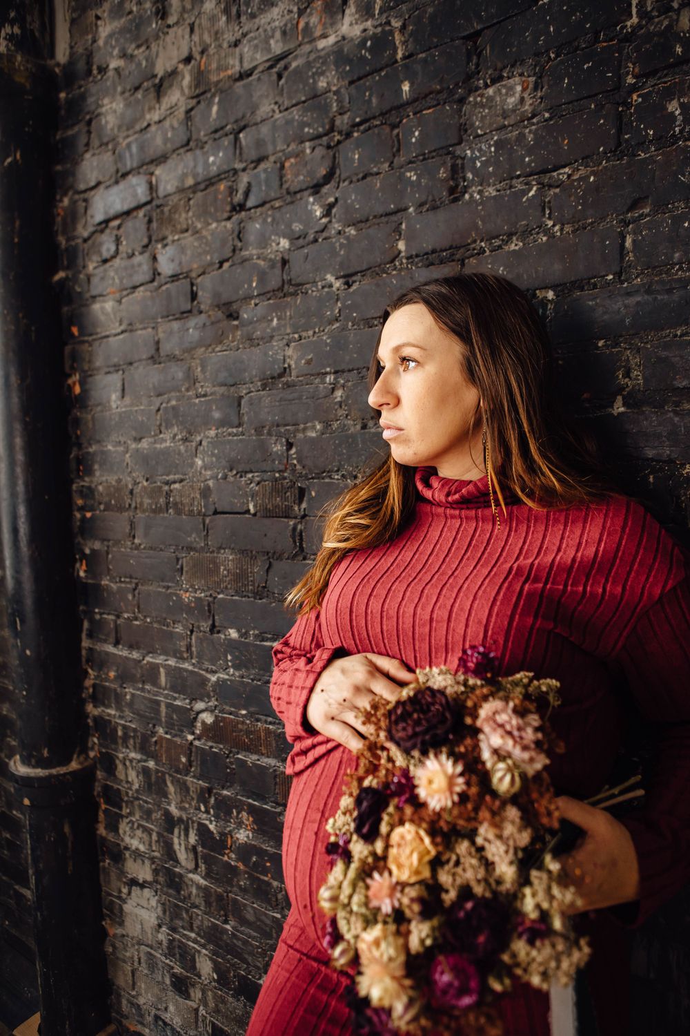 A woman in a red sweater holds dried flowers while standing against a dark brick wall in moody lighting.