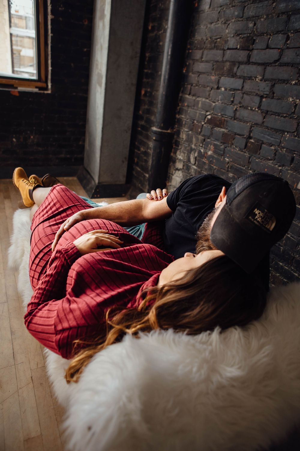 Person in plaid flannel and baseball cap relaxes on soft white fur rug against exposed brick wall interior.