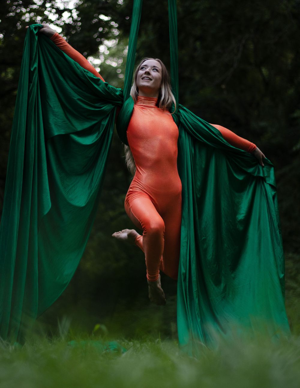 Performer in orange bodysuit poses dramatically with green silk aerial curtains in forest setting.