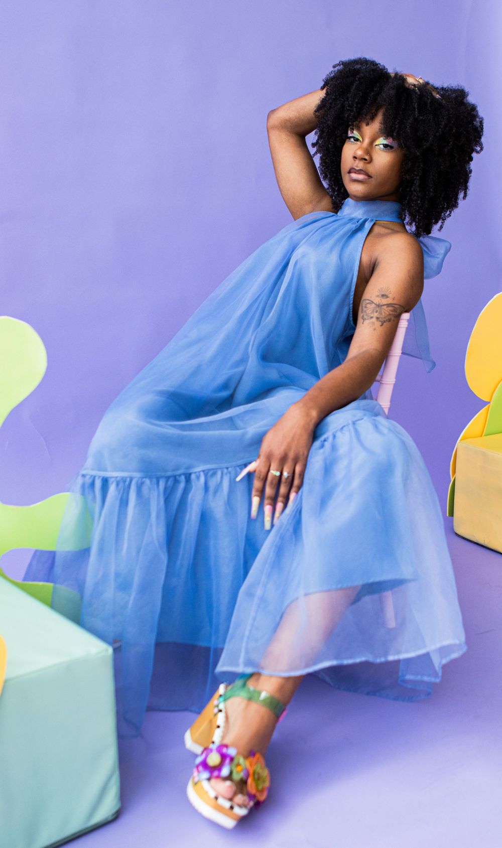 Fashion portrait in flowing blue tulle dress against purple backdrop with pastel geometric props.