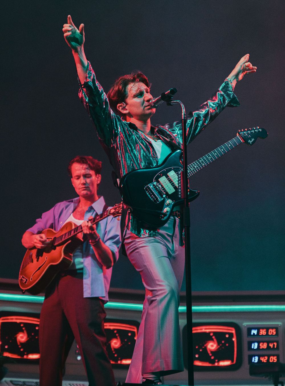 Performer in metallic jacket raises arms triumphantly on stage with guitarist.