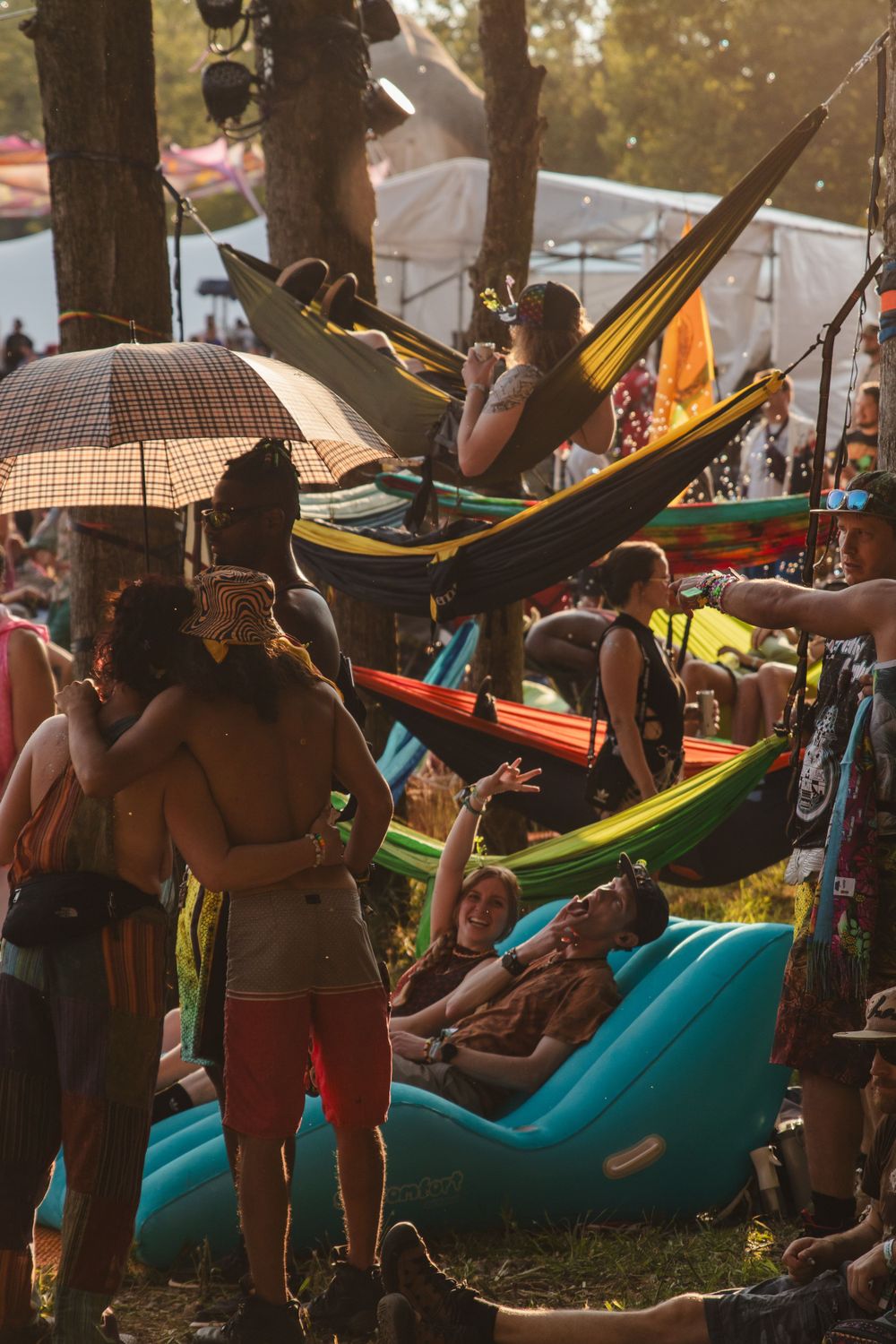 Festival goers relax in colorful hammocks under trees during sunset with umbrellas.