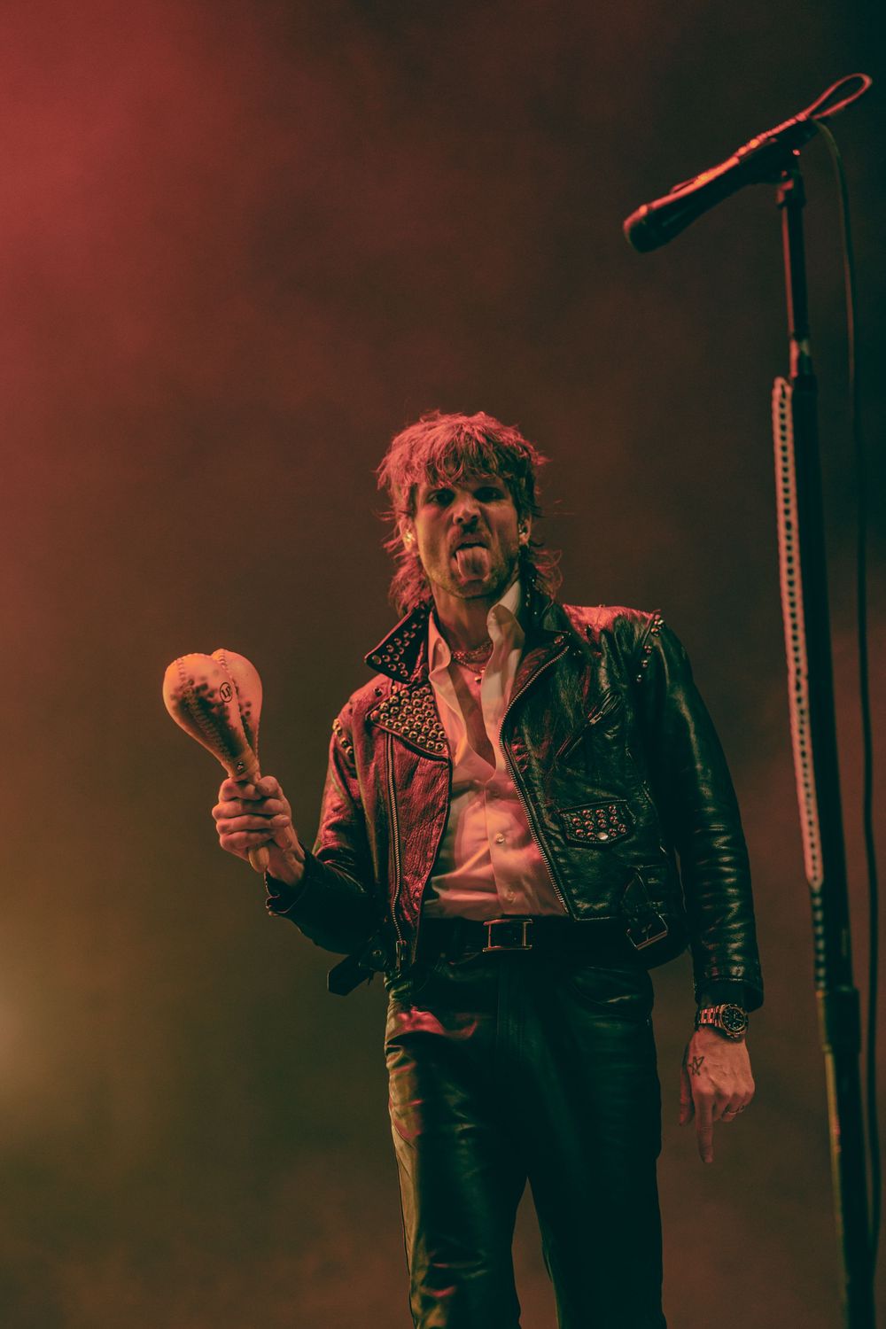 Rock performer in studded leather jacket holds a maraca on a smoky red-lit stage near a microphone stand.