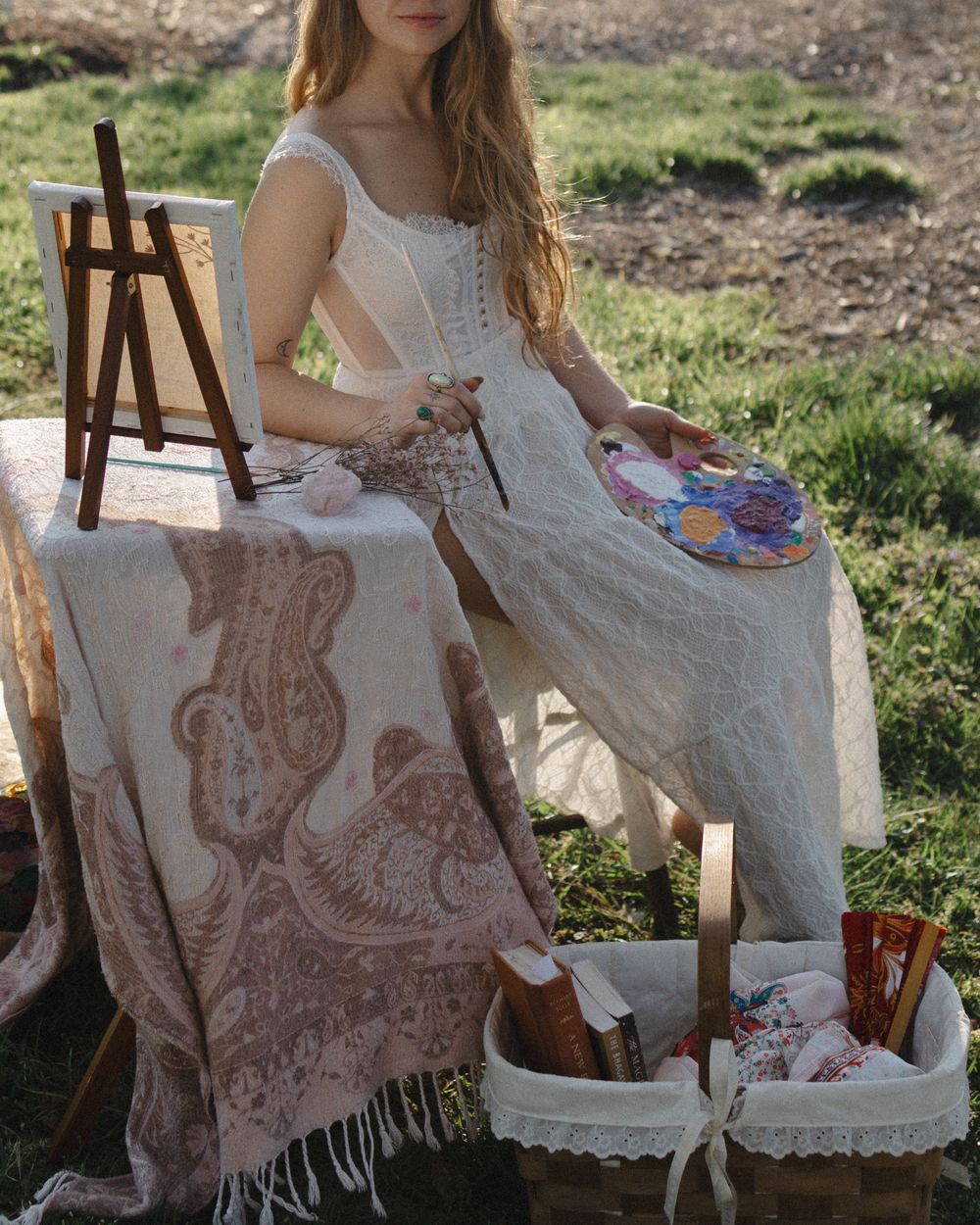 Woman in white lace dress painting outdoors with easel, palette, and art supplies basket on grass.